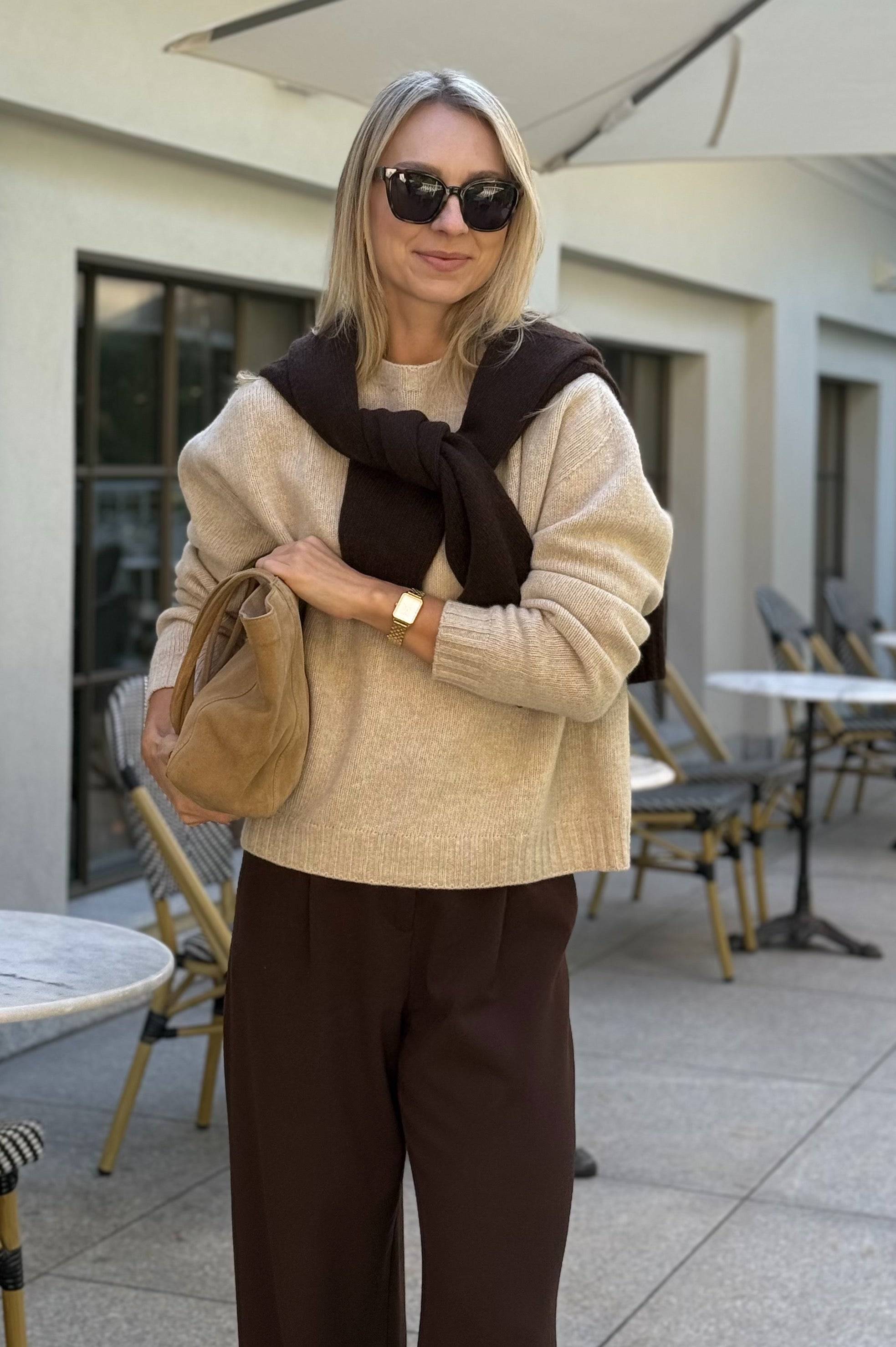Sweter Olmera beżowy, wełniany, brązowe spodnie eleganckie, stylizacja z torebką i kawą. Zarzucony na ramiona sweter Olmera w czekoladowym kolorze