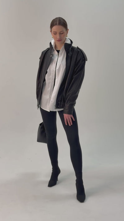 Czarna skórzana kurtka w stylu lat 90. oversize w stylizacji z rozpiętą białą koszulą, czarnymi spodniami skinny jeans i czarną torebką 